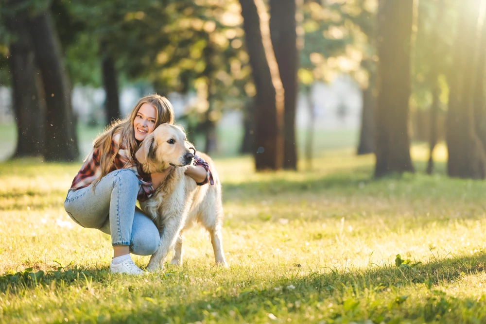 Smiling woman kneeling beside her Golden Retriever in a sunlit park, showcasing the bond built with top dog trainers.